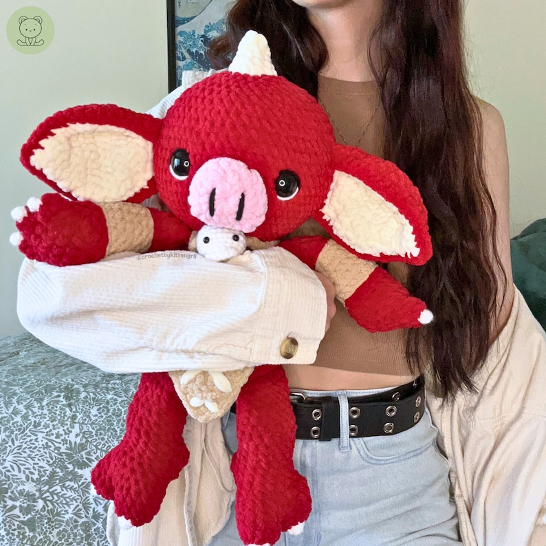 May include: A red and white crocheted plush toy shaped like a dragon with a pink nose and black eyes. The toy is being held by a person wearing a white shirt and blue jeans.