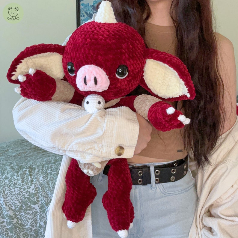 May include: A red and white crocheted plush toy shaped like a dragon with a pink nose and large eyes. The toy is being held by a person wearing a white corduroy jacket and a black belt.