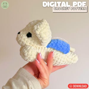 May include: A cream-coloured, crocheted dog toy with black eyes and a blue accent. The plush dog is lying down and held in a person's hand. The image includes text that reads "DIGITAL PDF CROCHET PATTERN".