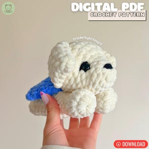 May include: A cream-coloured, handmade crochet dog with black eyes and a blue cape. The image includes the text "DIGITAL PDF CROCHET PATTERN" and "DOWNLOAD". The dog is held in a person's hand.