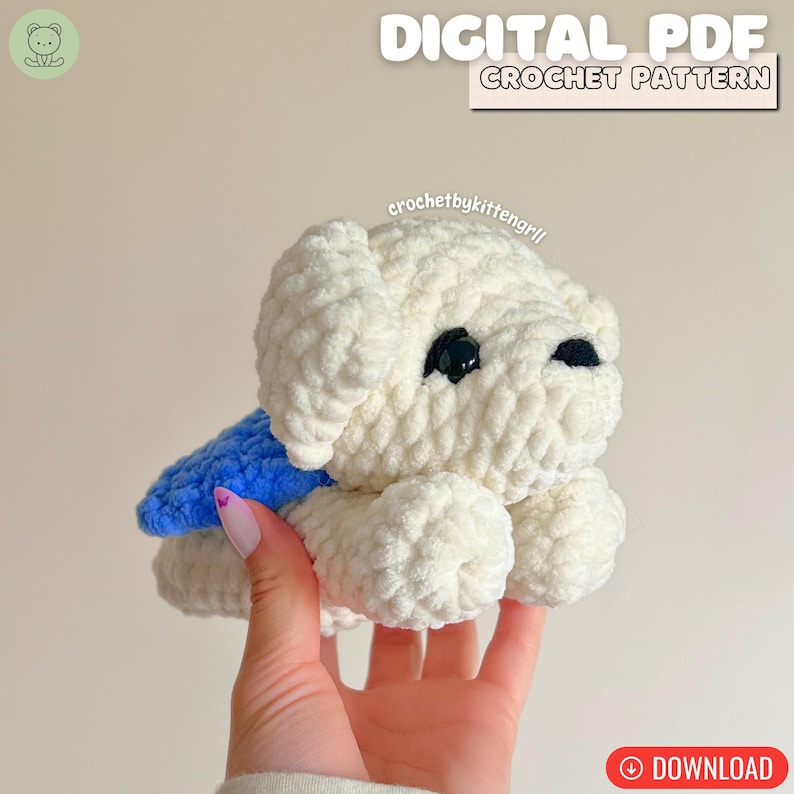 May include: A cream-coloured, handmade crochet dog with black eyes and a blue cape. The image includes the text "DIGITAL PDF CROCHET PATTERN" and "DOWNLOAD". The dog is held in a person's hand.
