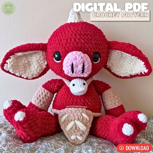 May include: A crocheted plush toy in the shape of a red creature with a horn, white ears, and a pink snout. The toy is wearing a brown crocheted belt and a white crocheted necklace. The toy is sitting on a patterned fabric.