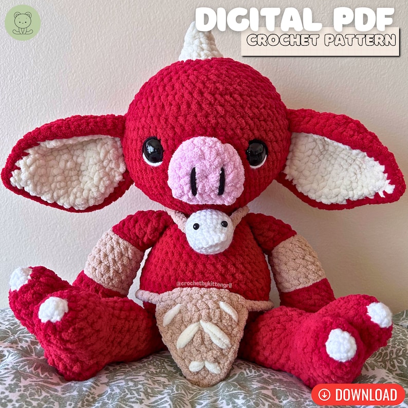 May include: A crocheted plush toy in the shape of a red creature with a horn, white ears, and a pink snout. The toy is wearing a brown crocheted belt and a white crocheted necklace. The toy is sitting on a patterned fabric.