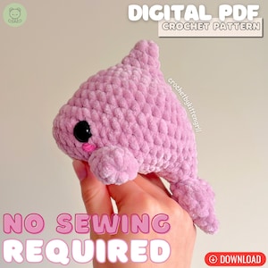 Op de afbeelding: Een lichtroze gehaakt pluche dolfijnspeeltje met grote zwarte veiligheidsogen en kleine roze geborduurde wangen. De tekst op de afbeelding luidt DIGITAL PDF CROCHET PATTERN, crochetbykittengrill en NO SEWING REQUIRED met downloadicoon.