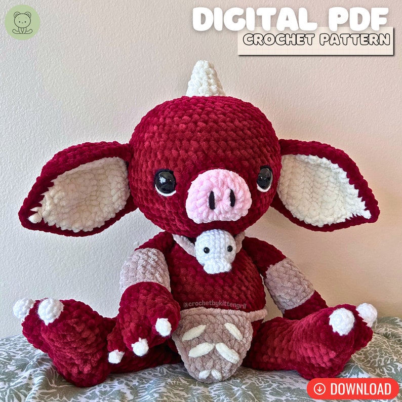 May include: Crochet pattern for a red and white plush toy with a horn, pig nose, and a small white plush toy around its neck. The pattern is available as a digital PDF download. "acrochetbykittengirl" is written on the toy's belly.