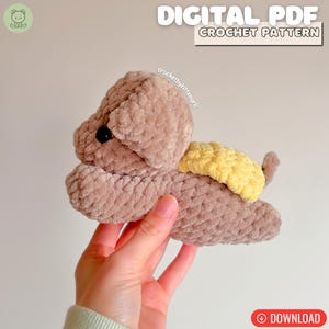 May include: A crocheted plush puppy in shades of brown and yellow. The puppy has a textured, soft appearance, with a yellow wing-like detail on its back. The image includes the text "DIGITAL PDF CROCHET PATTERN" and a download button.
