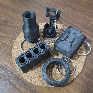 May include: A collection of five black 3D-printed automotive-themed keychains, including a socket, a piston and connecting rod, a fuel canister, an engine block, and a tire, each attached to a silver metal split ring for keys.