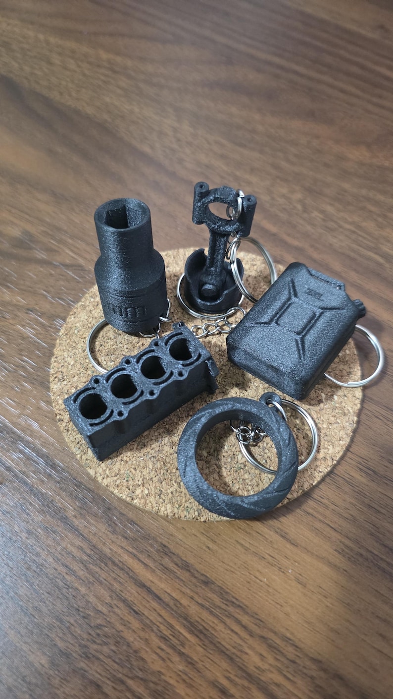 May include: A collection of five black 3D-printed automotive-themed keychains, including a socket, a piston and connecting rod, a fuel canister, an engine block, and a tire, each attached to a silver metal split ring for keys.