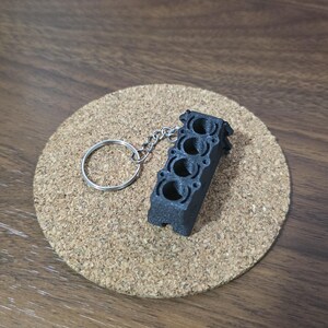 May include: A black, 3D-printed keychain shaped like a four-cylinder engine block. The object features four distinct circular cylinder bores arranged in a straight line and is attached to a standard silver-colored metal split ring key holder.