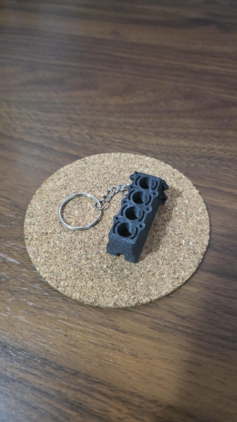 May include: A black, 3D-printed keychain shaped like a four-cylinder engine block. The object features four distinct circular cylinder bores arranged in a straight line and is attached to a standard silver-colored metal split ring key holder.