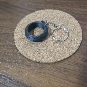 May include: A black keychain featuring a circular tire-shaped charm with a textured tread pattern, attached to a silver-colored metal chain and a round split ring for keys. The object is presented as a single unit with a durable matte finish.