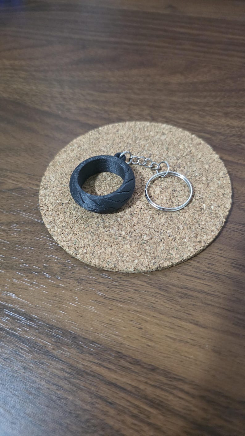 May include: A black keychain featuring a circular tire-shaped charm with a textured tread pattern, attached to a silver-colored metal chain and a round split ring for keys. The object is presented as a single unit with a durable matte finish.
