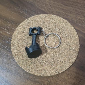 May include: A black keychain featuring a miniature engine piston and connecting rod assembly attached to a circular silver metal split ring. The object is constructed with a matte finish and displays the mechanical components of a combustion engine.