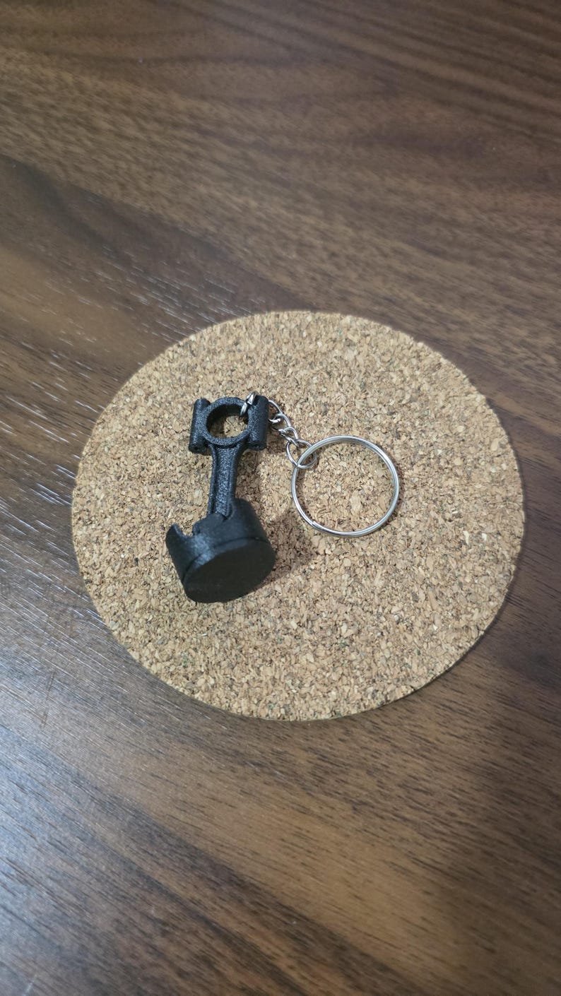 May include: A black keychain featuring a miniature engine piston and connecting rod assembly attached to a circular silver metal split ring. The object is constructed with a matte finish and displays the mechanical components of a combustion engine.