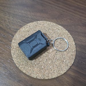 May include: A black miniature fuel canister keychain featuring a textured surface and a classic jerry can design. The small rectangular container is attached to a circular silver metal split ring, providing a functional accessory for keys.