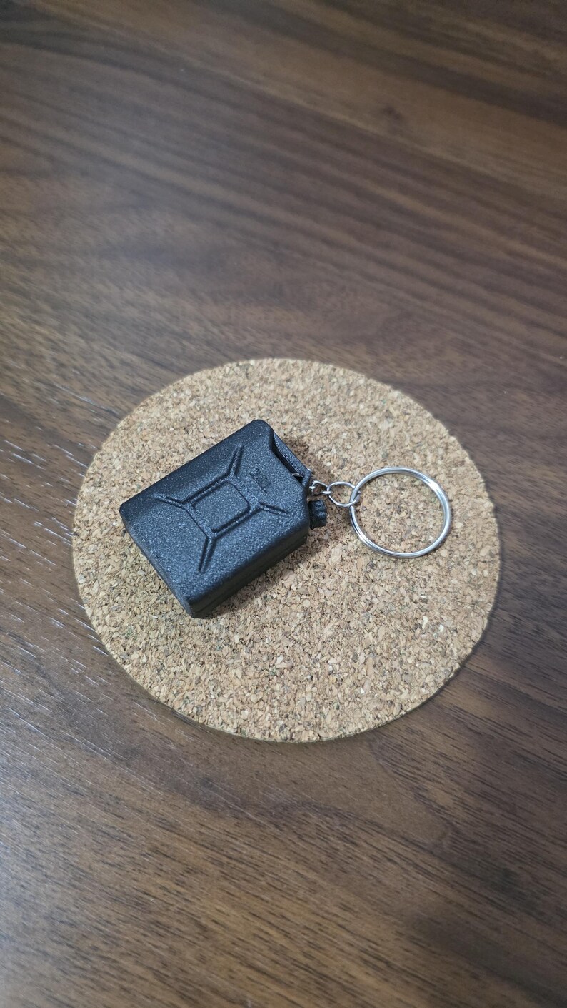 May include: A black miniature fuel canister keychain featuring a textured surface and a classic jerry can design. The small rectangular container is attached to a circular silver metal split ring, providing a functional accessory for keys.