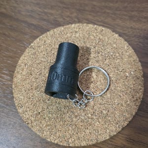 May include: A black, textured, cylindrical socket keychain with the text 10mm embossed on the side. The socket is attached to a silver-colored metal split ring by a short, thin metal chain, creating a functional tool-themed accessory.