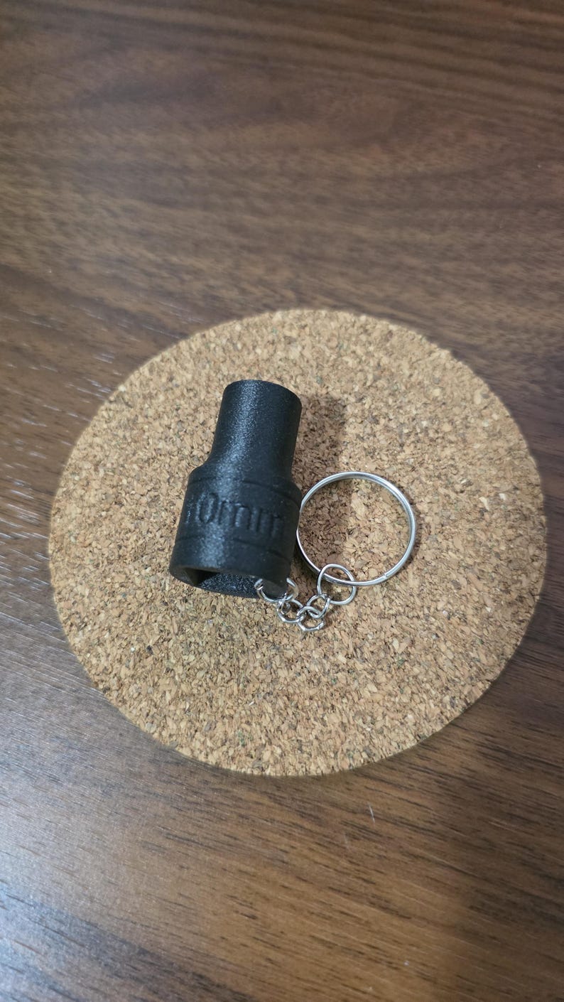 May include: A black, textured, cylindrical socket keychain with the text 10mm embossed on the side. The socket is attached to a silver-colored metal split ring by a short, thin metal chain, creating a functional tool-themed accessory.