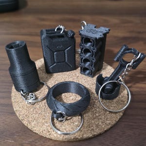 May include: A collection of five black 3D-printed keychains featuring automotive-themed designs, including a fuel canister, an engine block, a piston with a connecting rod, a tire-shaped ring, and a cylindrical socket tool, all with metal rings.