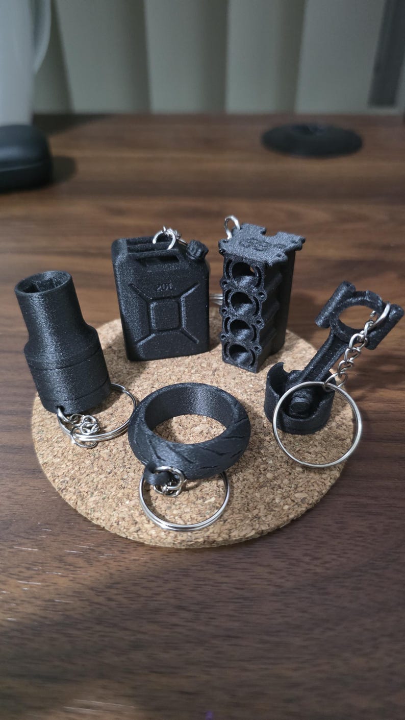 May include: A collection of five black 3D-printed keychains featuring automotive-themed designs, including a fuel canister, an engine block, a piston with a connecting rod, a tire-shaped ring, and a cylindrical socket tool, all with metal rings.