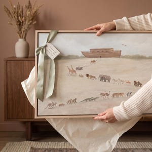 May include: A rectangular framed wall art print featuring a painted scene of a wooden ark on a hill with various animals walking in pairs toward it. A sage green satin ribbon is tied to the top left corner with a small white paper gift tag attached.