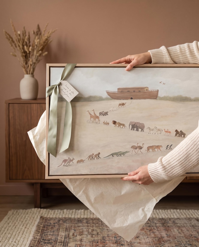 May include: A rectangular framed wall art print featuring a painted scene of a wooden ark on a hill with various animals walking in pairs toward it. A sage green satin ribbon is tied to the top left corner with a small white paper gift tag attached.
