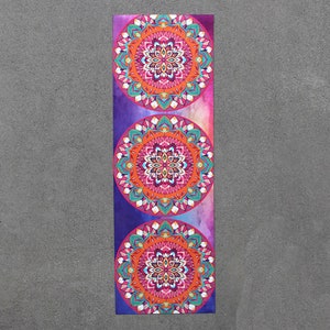 May include: A yoga mat with a colorful mandala design. The mat features three large, intricate mandalas in shades of pink, orange, blue, and green. The background is a watercolor-like purple and pink.