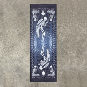 May include: A blue yoga mat with a white design of two skeletons with wings, hearts, and a mandala pattern.