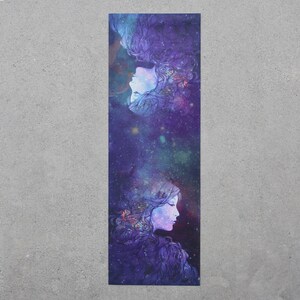 May include: A long, narrow skateboard deck with a colorful design featuring two women's faces with flowing hair and a galaxy background. The design is in shades of purple, blue, and white.