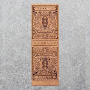 May include: A brown yoga mat with a decorative design featuring a stylized face and the word "Namaste" printed on it. The design also includes the text "The Light in Me Recognizes the Light in You".