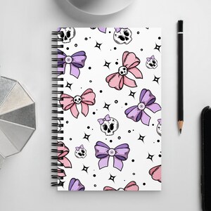 May include: A white spiral notebook with a repeating pattern of pink and purple bows and black skulls. The notebook has a white background with black stars.