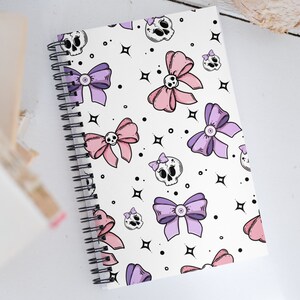 May include: A white spiral notebook with a pink and purple bow and skull pattern. The notebook has a white background with black outlines.