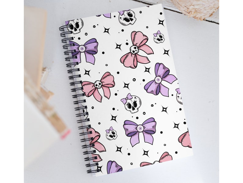 May include: A white spiral notebook with a pink and purple bow and skull pattern. The notebook has a white background with black outlines.