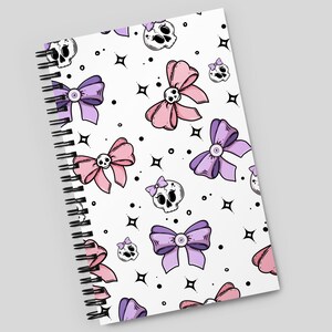 May include: A white spiral notebook with a pink and purple bow and skull pattern. The notebook has a white background with black outlines.