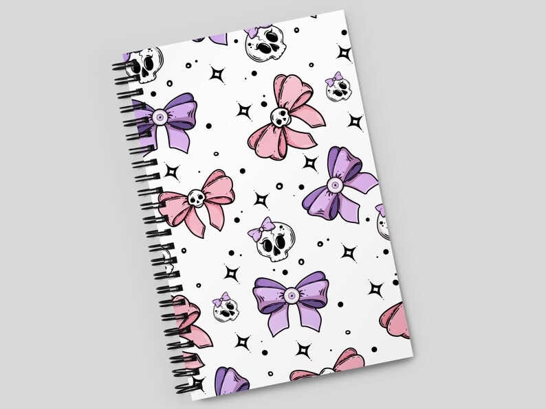 May include: A white spiral notebook with a pink and purple bow and skull pattern. The notebook has a white background with black outlines.