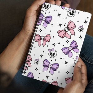 May include: A white spiral notebook with a pink and purple bow and skull pattern. The notebook has a white background with black outlines.