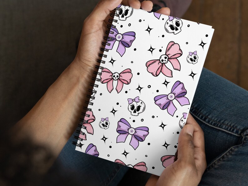 May include: A white spiral notebook with a pink and purple bow and skull pattern. The notebook has a white background with black outlines.