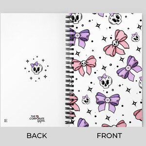 May include: A white spiral notebook with a pink and purple skull and bow pattern on the front cover. The back cover is white with the text "THE CORPORATE GOTH." printed in black.