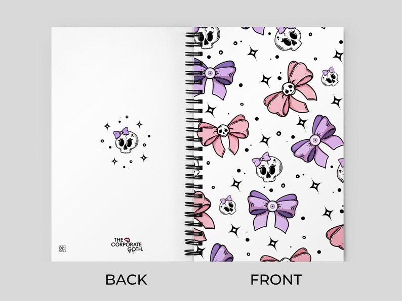 May include: A white spiral notebook with a pink and purple skull and bow pattern on the front cover. The back cover is white with the text "THE CORPORATE GOTH." printed in black.