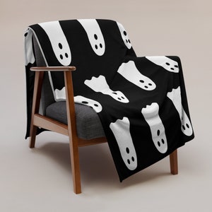 May include: A black and white throw blanket with a repeating pattern of white ghosts. The blanket is draped over a wooden chair.