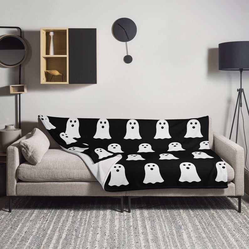 May include: A black and white throw blanket with a repeating pattern of white ghosts. The blanket is draped over a gray couch.