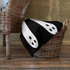 May include: A black and white throw blanket with a pattern of two white ghosts with black eyes. The blanket is draped over a wicker basket.