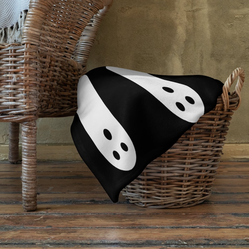 May include: A black and white throw blanket with a pattern of two white ghosts with black eyes. The blanket is draped over a wicker basket.