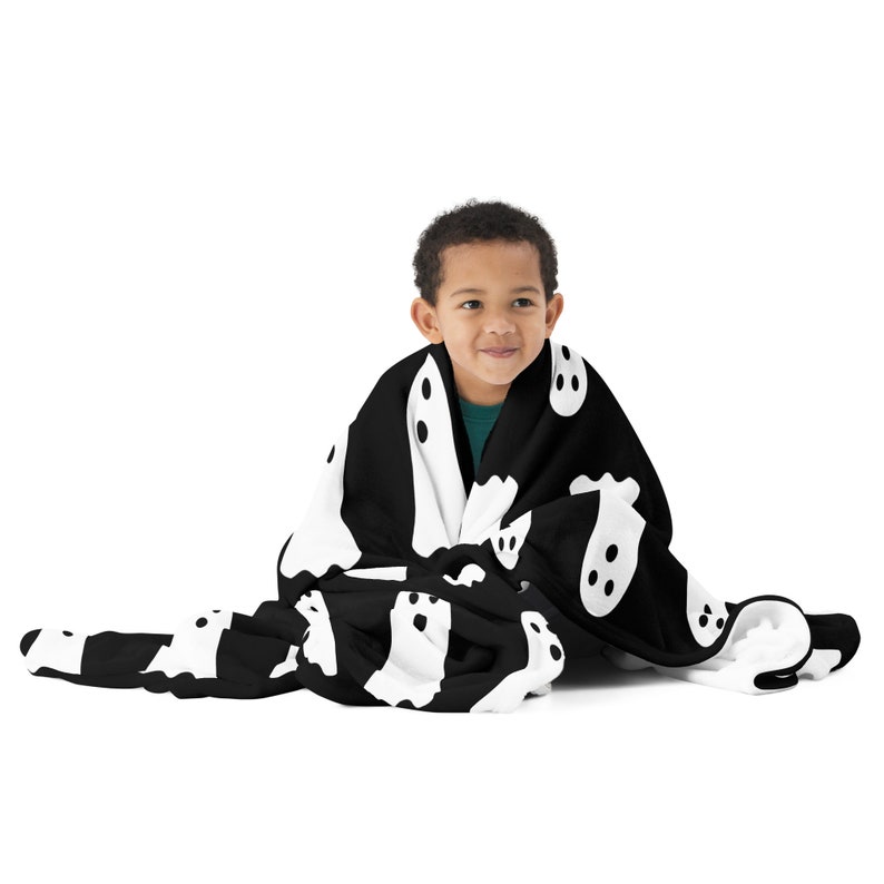May include: A child is wrapped in a black and white fleece blanket with a ghost pattern. The blanket is soft and cozy, perfect for cuddling up on a cold night.