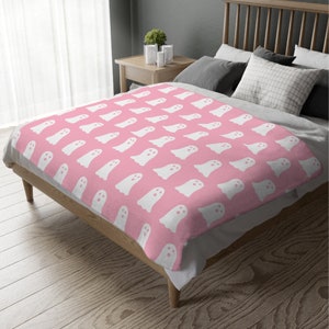 May include: A pink fleece throw blanket with a white ghost pattern. The blanket is folded and displayed on a bed.