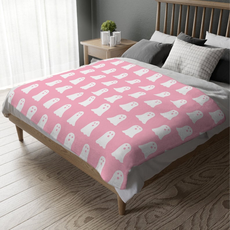 May include: A pink fleece throw blanket with a white ghost pattern. The blanket is folded and displayed on a bed.