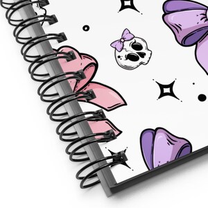 May include: A spiral bound notebook with a white cover featuring a repeating pattern of purple and pink bows, skulls with pink bows, and black stars and dots.