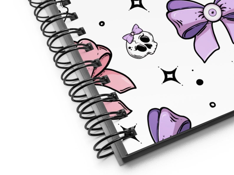 May include: A spiral bound notebook with a white cover featuring a repeating pattern of purple and pink bows, skulls with pink bows, and black stars and dots.