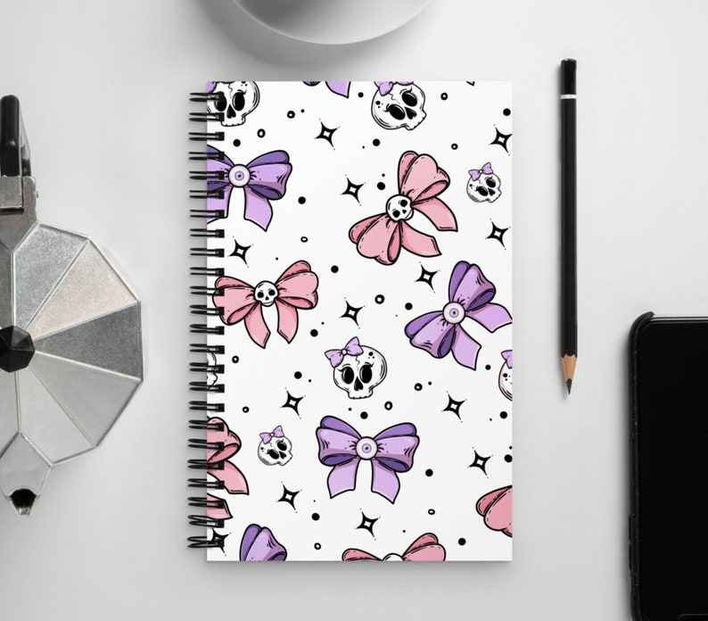 May include: A white spiral notebook with a repeating pattern of pink and purple bows and black skulls. The notebook has a white background with black stars.