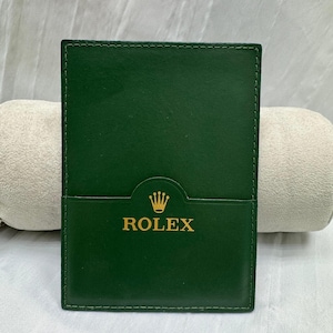 May include: A dark green leather wallet with the gold-colored word "ROLEX" and a crown logo. The wallet is rectangular with a pocket on the front and stitched edges. The background is a light-colored fabric.